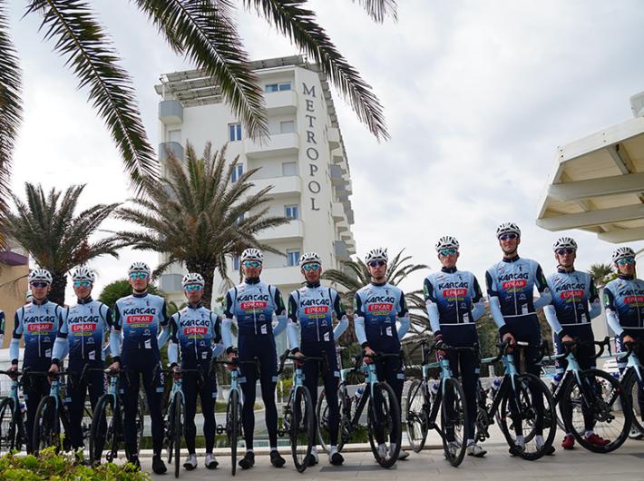 Ciclisti in divisa, pronti per la gara, davanti a un hotel e palme.