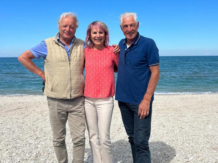 Drei lächelnde Personen umarmen sich an einem sonnigen Strand.